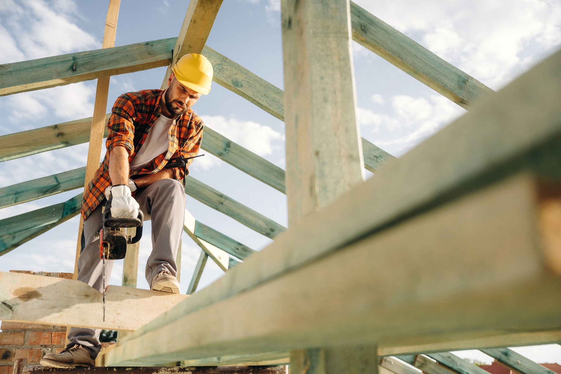 Looks down, cutting the plank with chainsaw. Construction worker is on the roof of a house